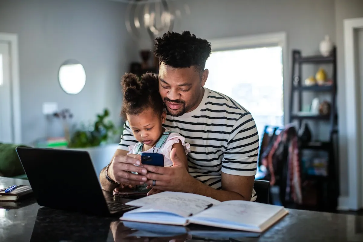 A father looks at his smart phone while his young daughter, sitting on his lap, plays with her own phone.