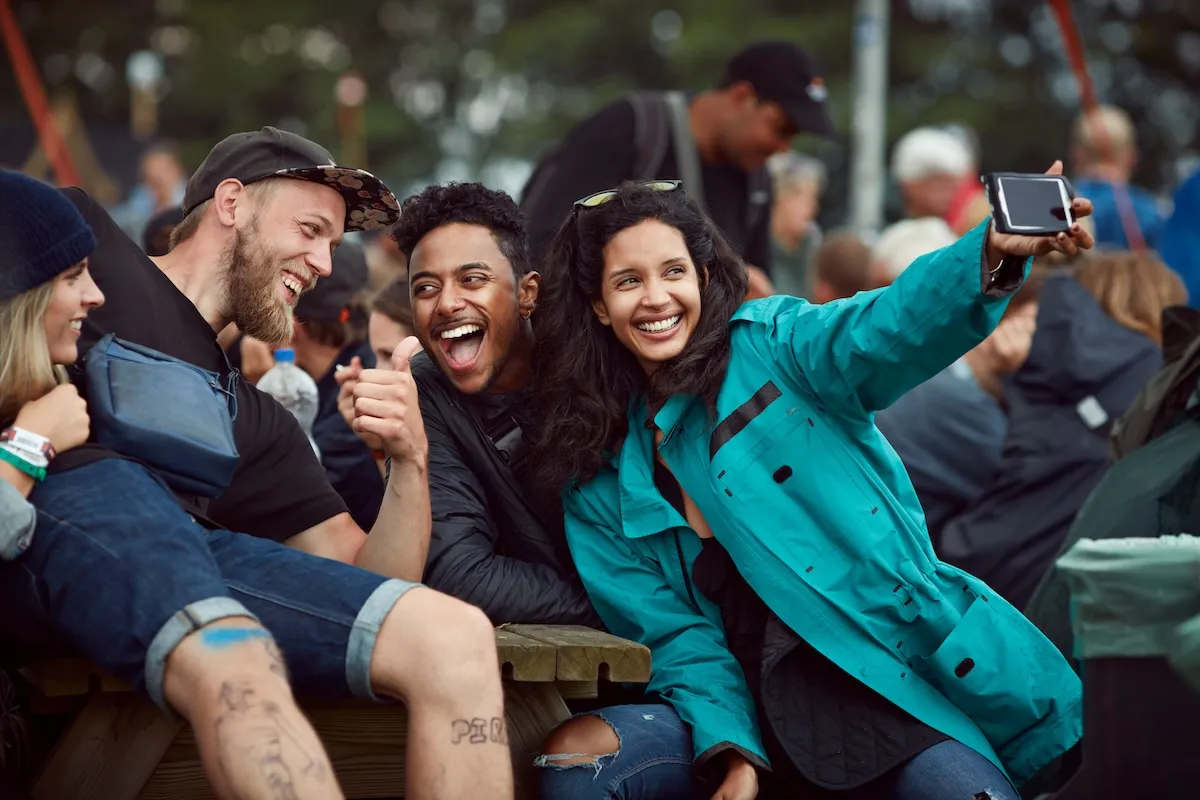 Four young friends laugh as they take a selfie on a mobile phonen in a busy park outside
