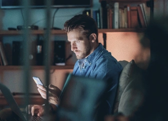 A person using a laptop and mobile phone at home, representing secure online activity protected by built‑in broadband security.